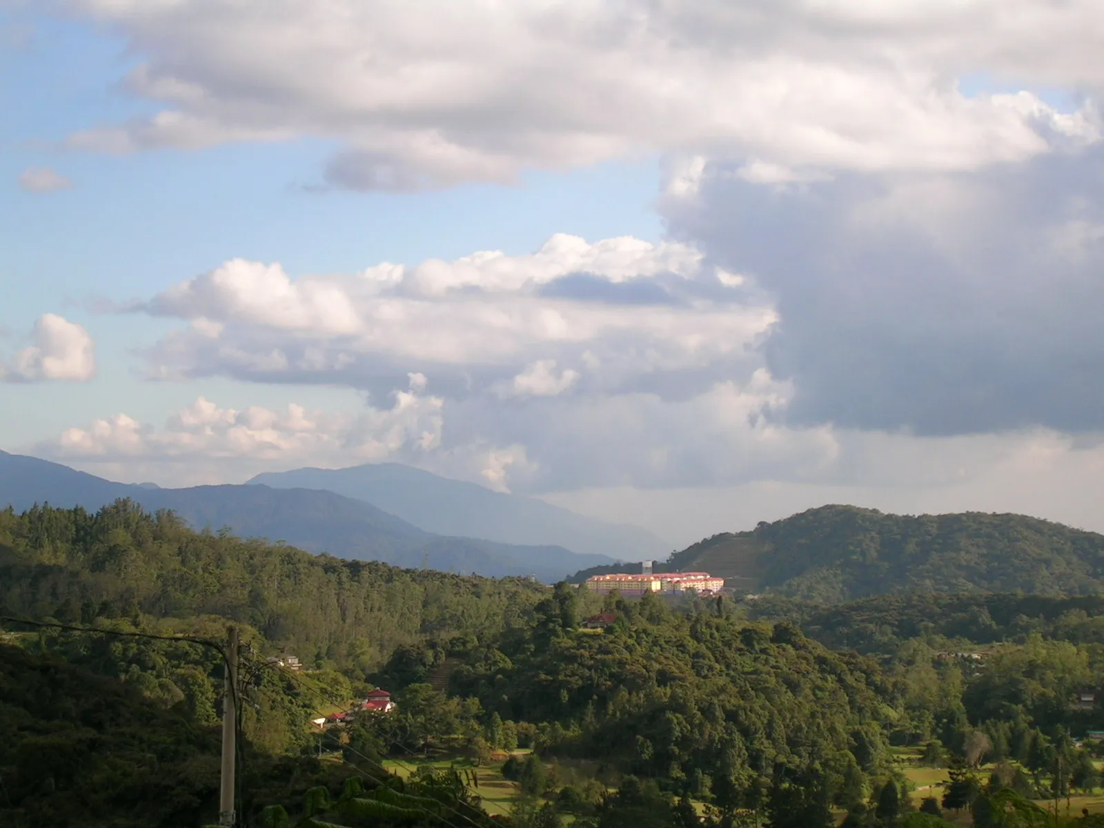 Forested hills with a large hotel building on a ridge, mountains and clouds behind